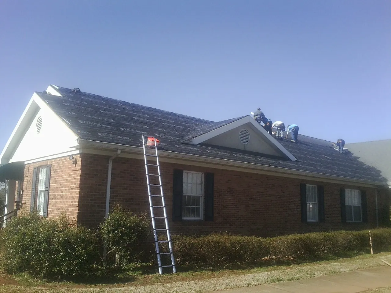 Crew tearing shingles off a brick ranch roof with a ladder set — Emergency Roof Repair work by our crew in Markesan