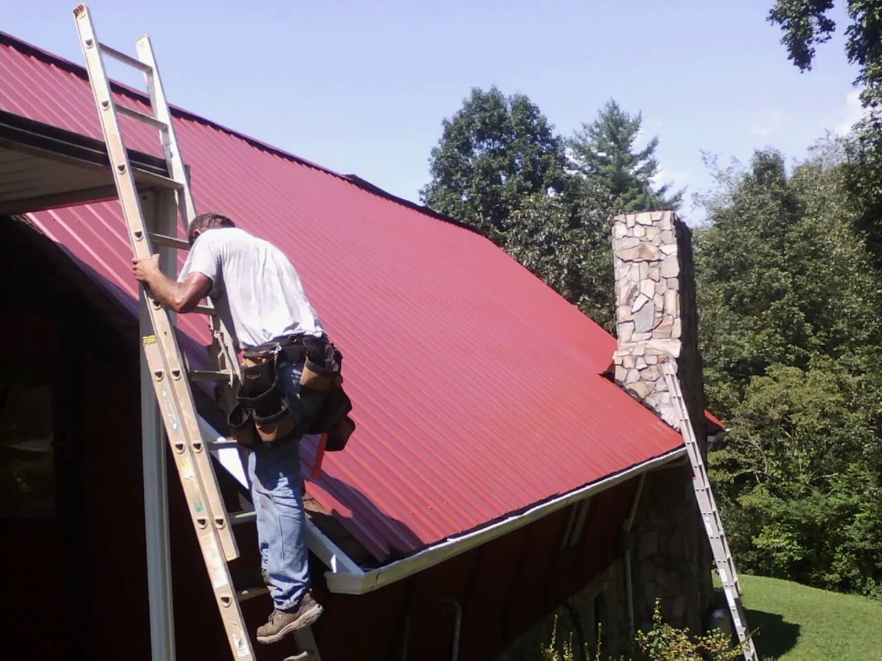 Roofer on a ladder beside a red metal roof and stone chimney — Soffit & Fascia Repair by Roofing Brothers in Markesan